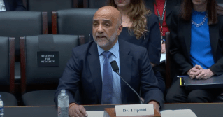 Micky Tripathi pictured sitting at a desk as a witness for a hearing on Capitol Hill. He wears a suit and has a name placard in front of him that reads "Dr. Tripathi."